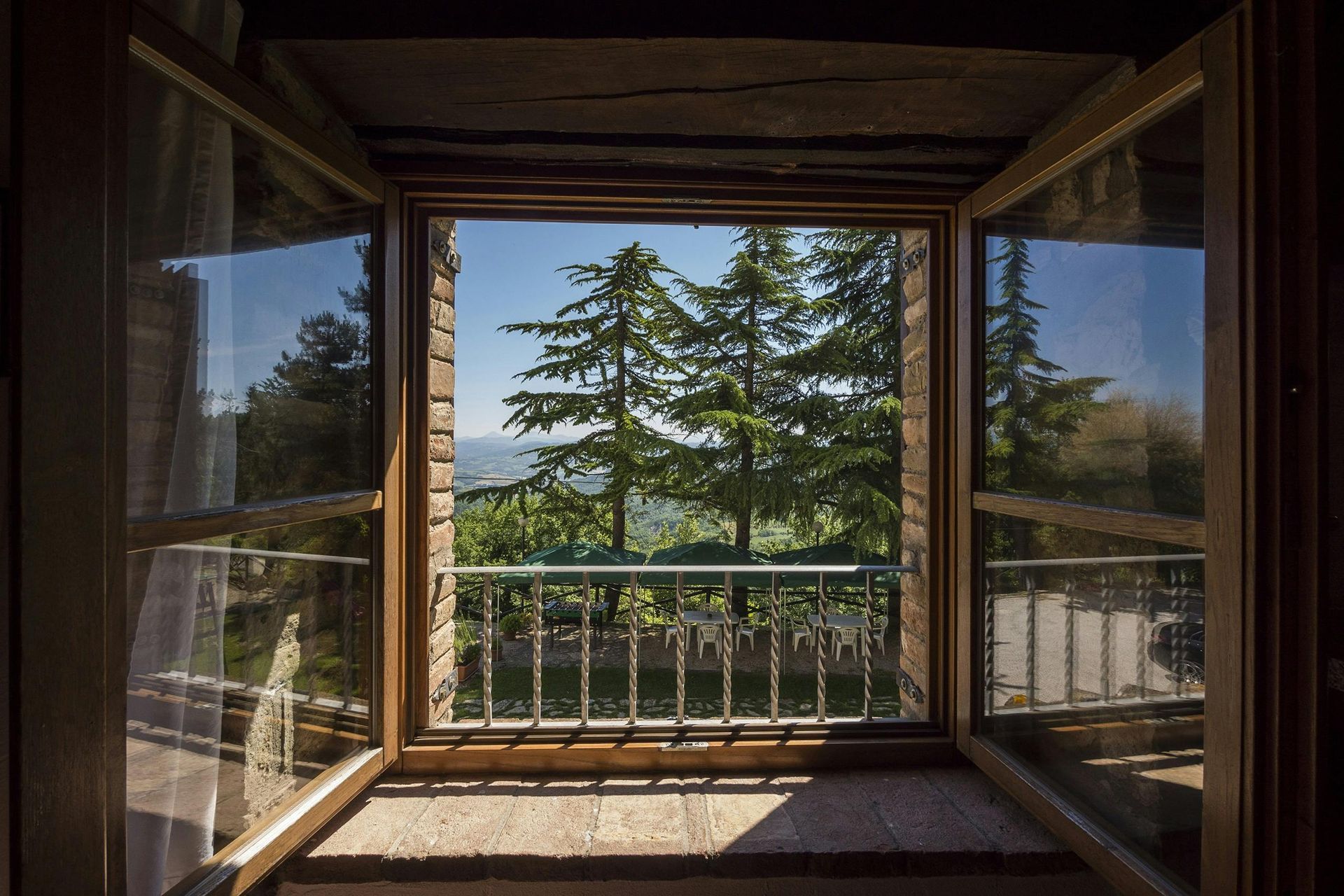 Open wooden window frames a view of tall green trees, a small balcony with a wrought-iron railing, and a clear blue sky.