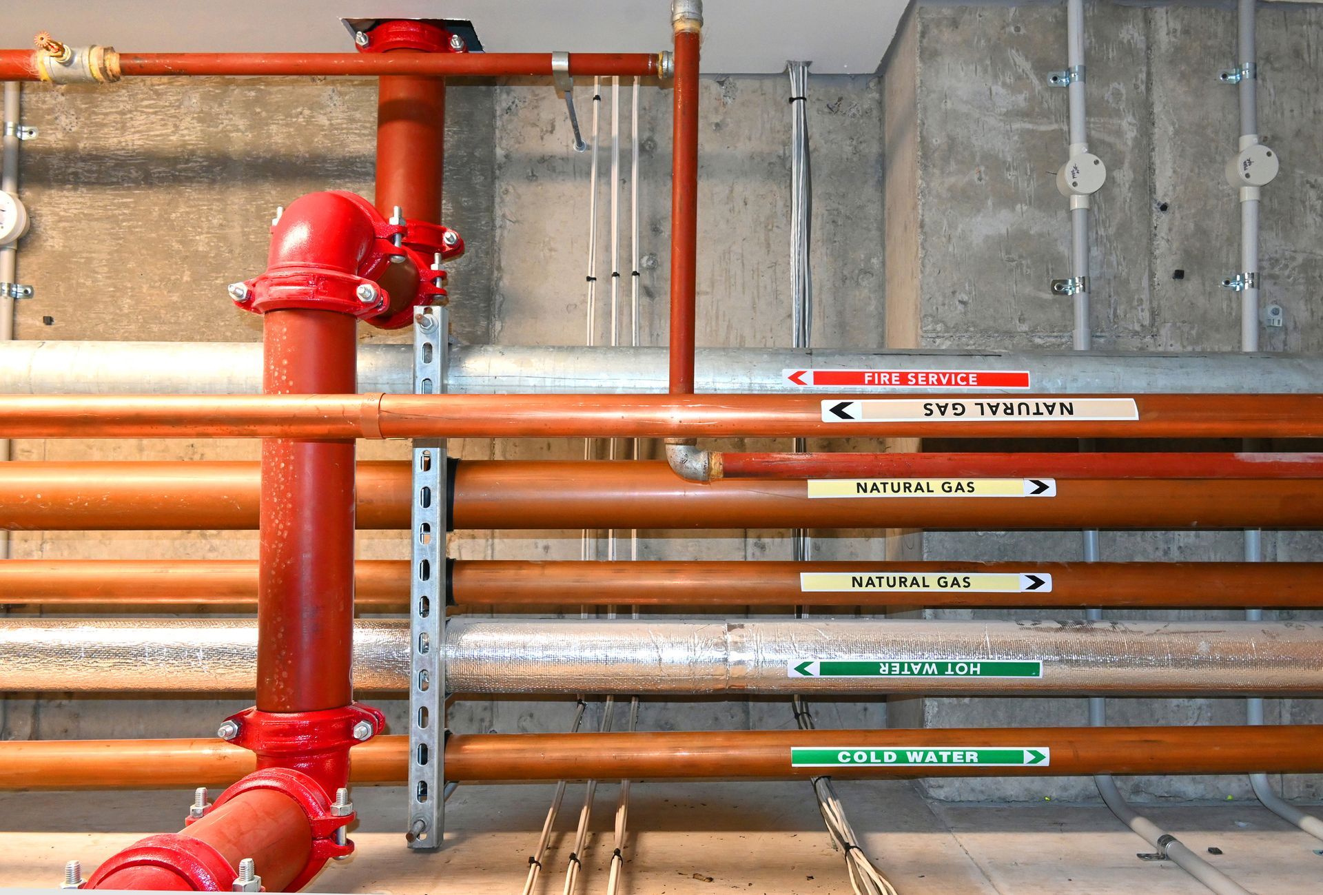 Red and copper pipes with labels, mounted on a concrete wall, likely for fire suppression and utilities.