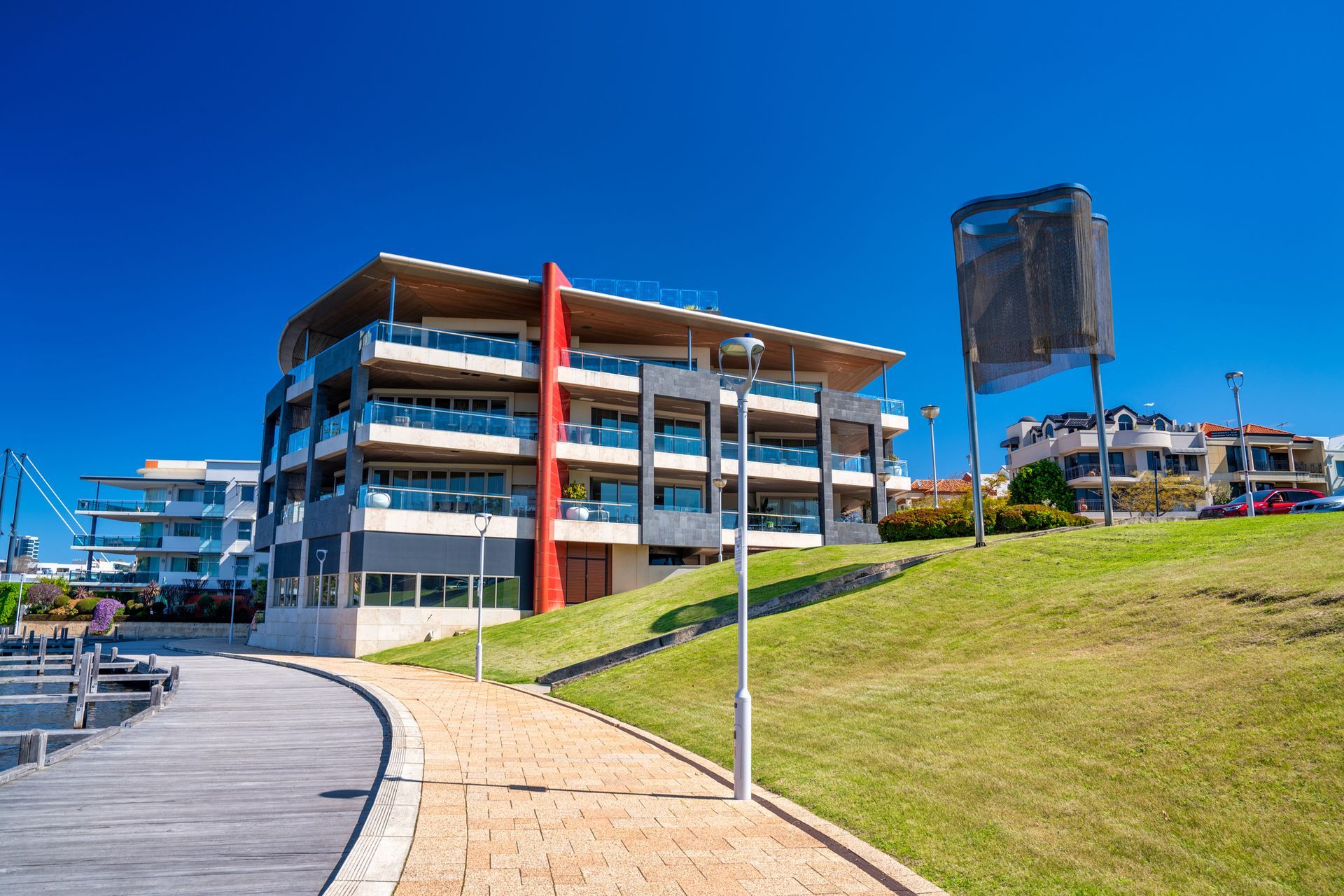Modern apartment building on a grassy hill next to a boardwalk, under a clear blue sky.