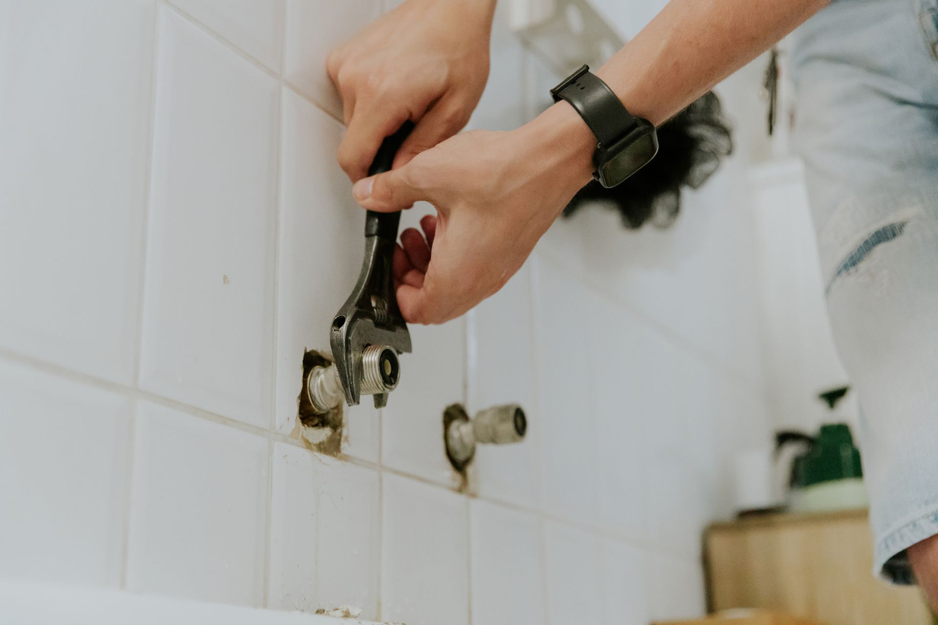 Person using a wrench on a pipe in a tiled bathroom.