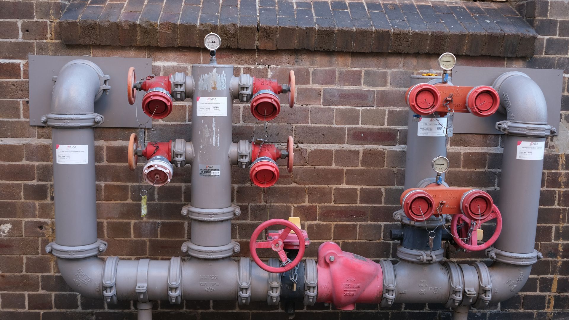 Gray fire hydrant system with red outlets and valves on a brick wall.