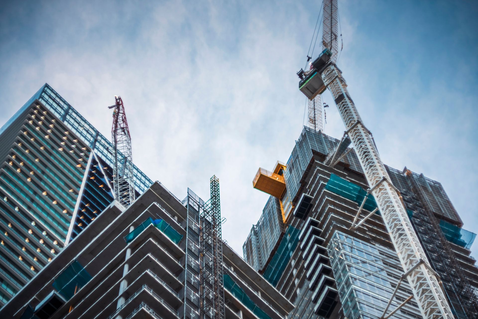 Construction cranes working on tall skyscrapers against a cloudy sky.