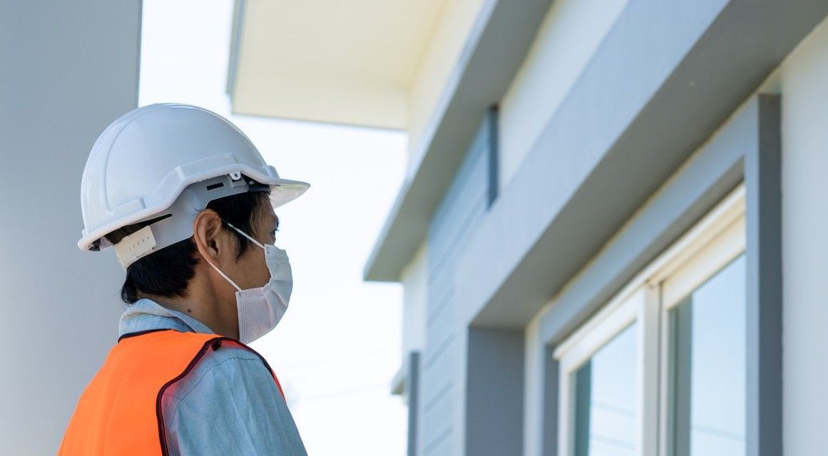 A man wearing a hard hat and a mask is standing in front of a building.