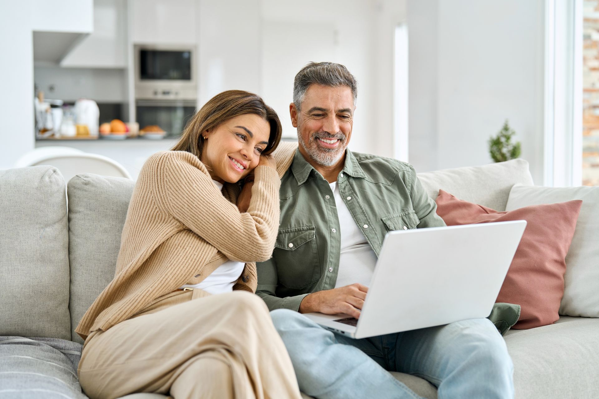 Couple on sofa, looking at laptop screen, smiling. Interior setting, natural light.