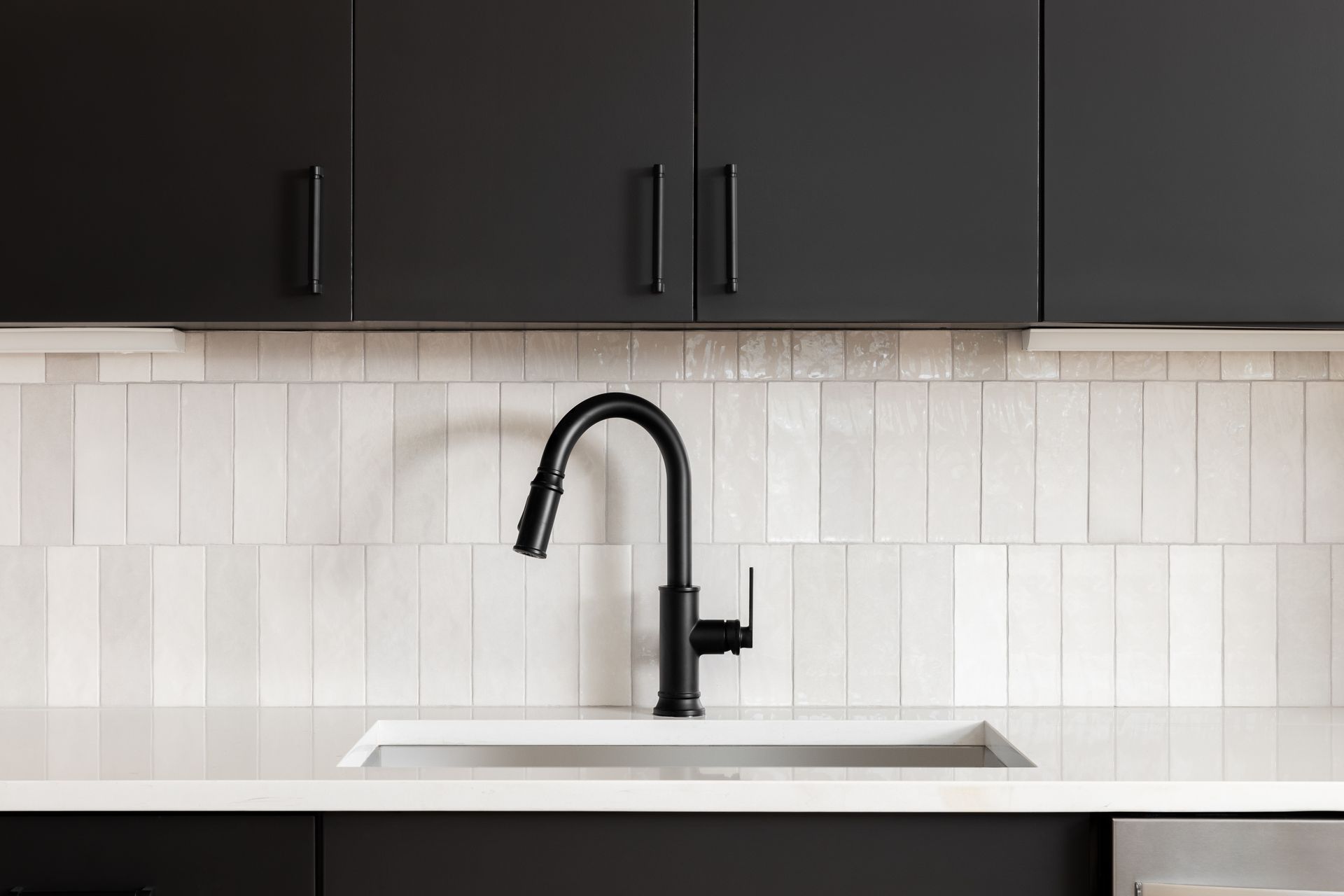 Black kitchen faucet over white sink and counter, with black cabinets and white tile backsplash.