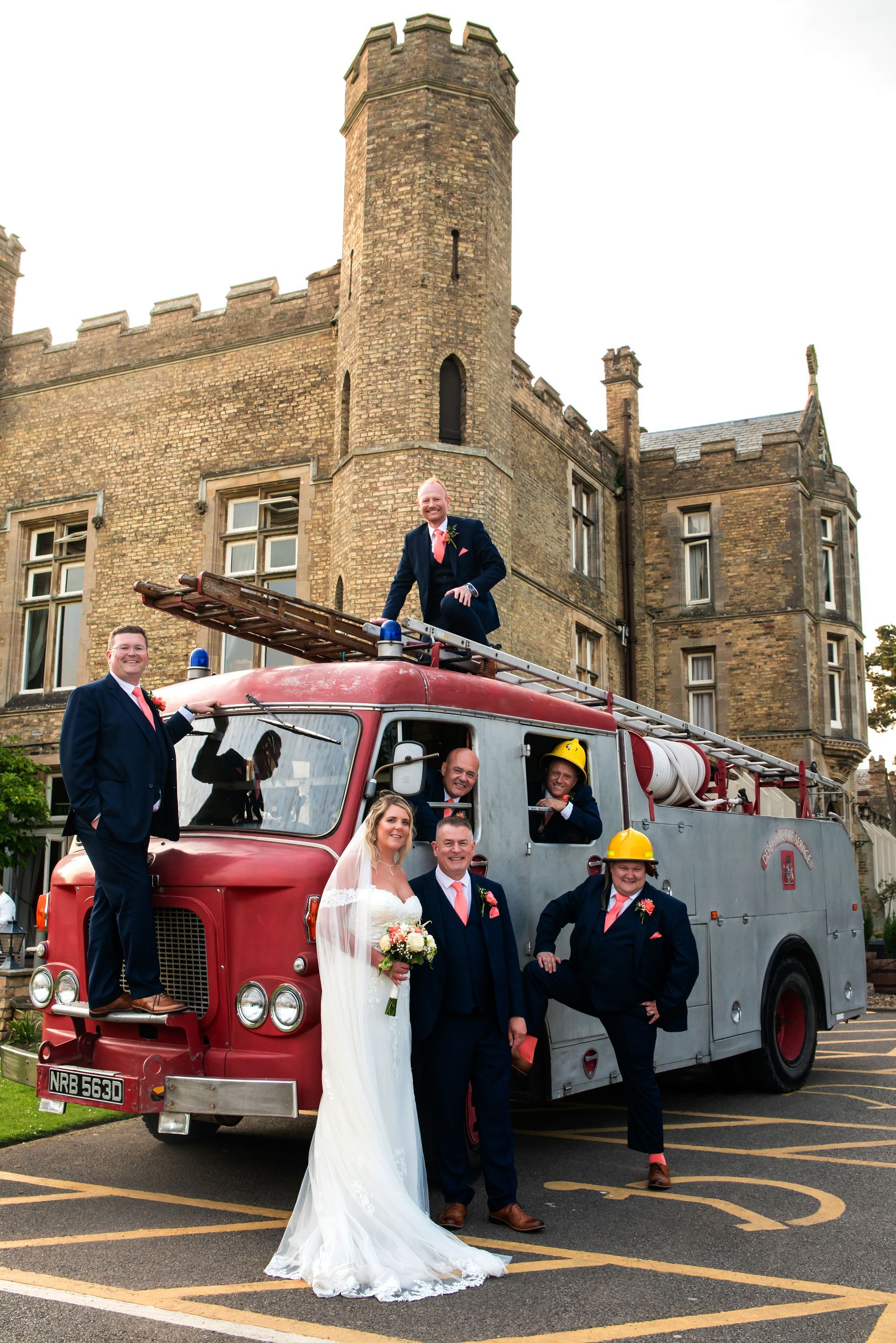 Bride and groom pose with friends on a vintage red fire truck in front of a stone building.