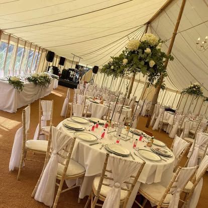 Wedding reception tent with round tables, white linens, floral centerpieces, and decorated chairs.