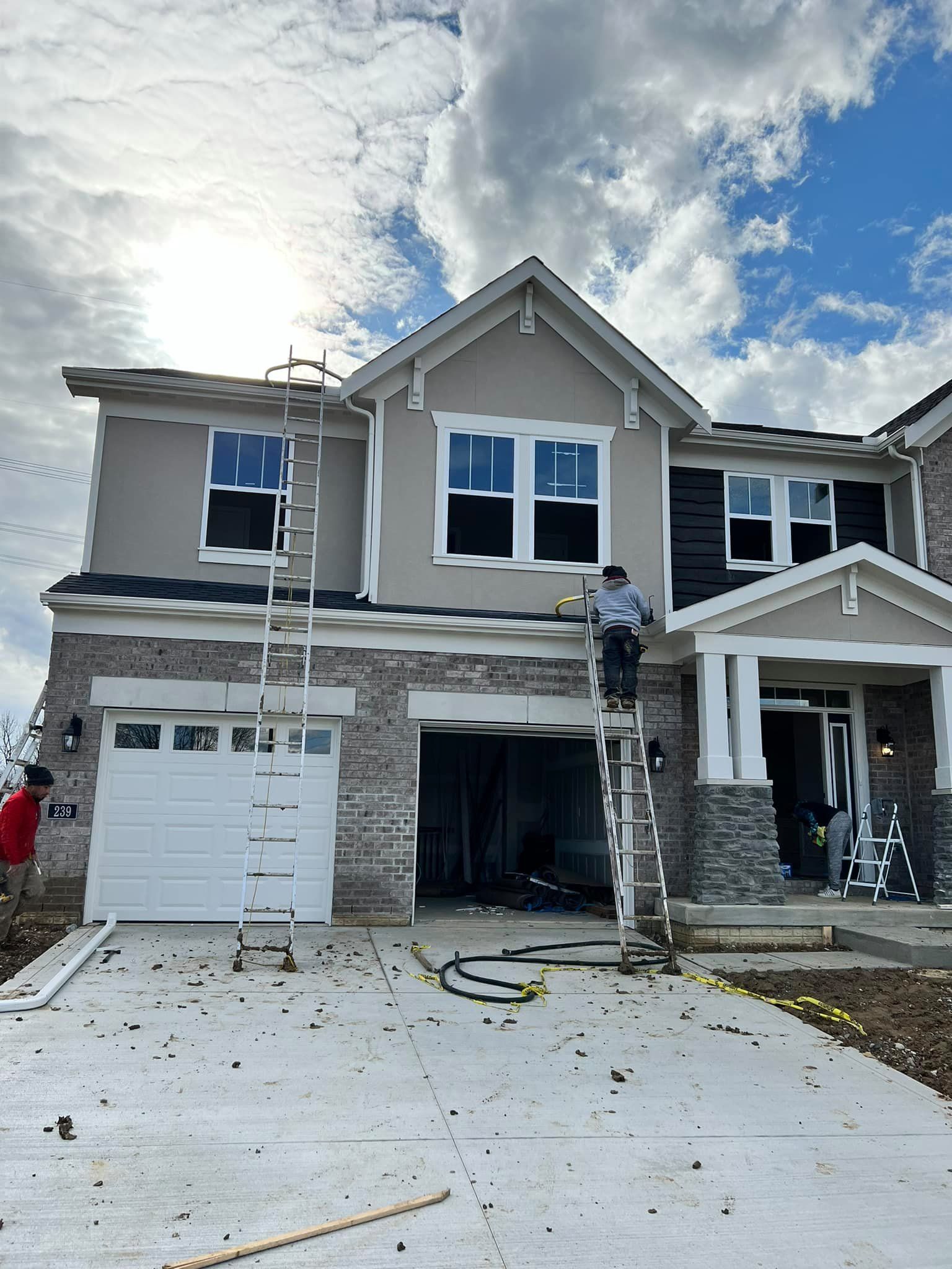 Two-story house under construction. Workers on ladders; grey exterior, white trim, garage. Cloudy sky.