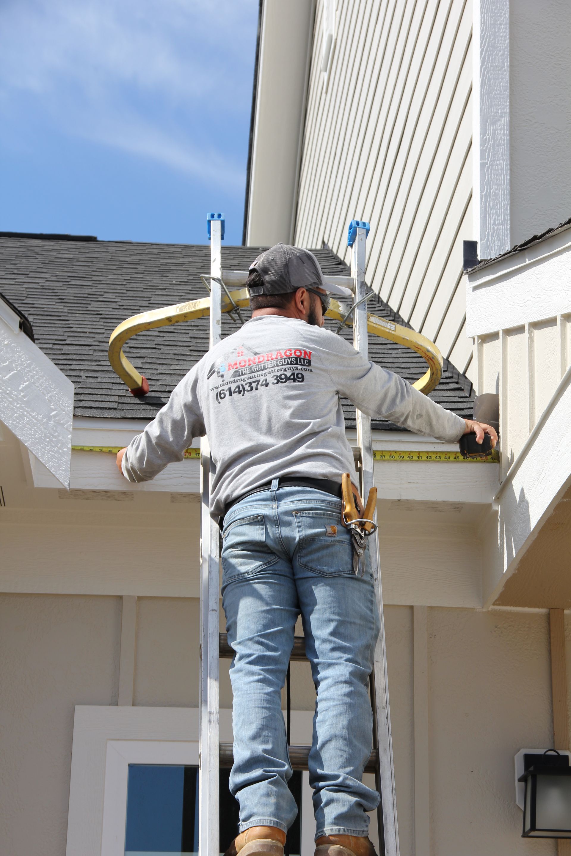 Man on ladder measuring eaves of a house with a tape measure, measuring for the installation of a gutter.