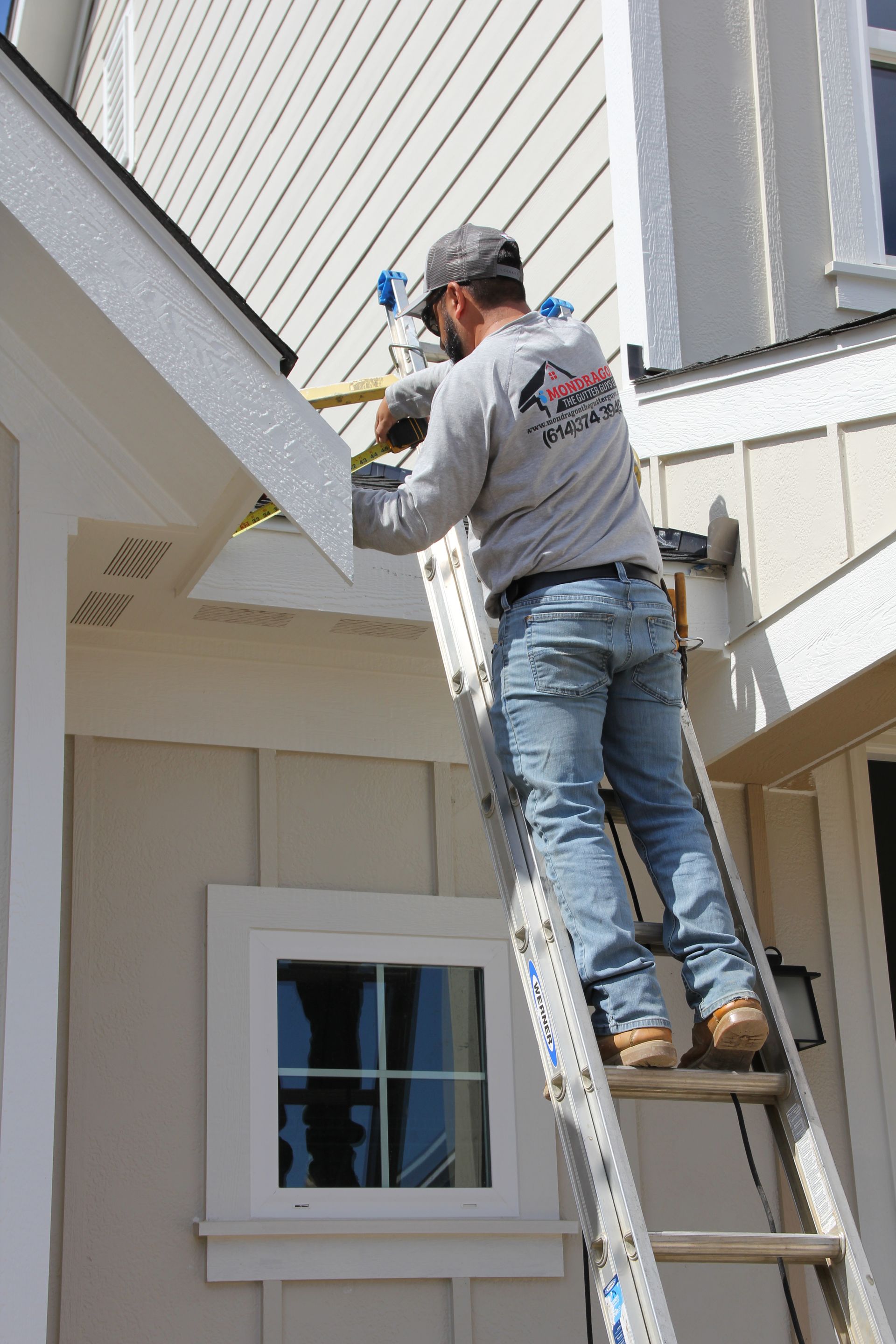 Man on ladder working on the side of a house; beige siding, white trim, gray sky.