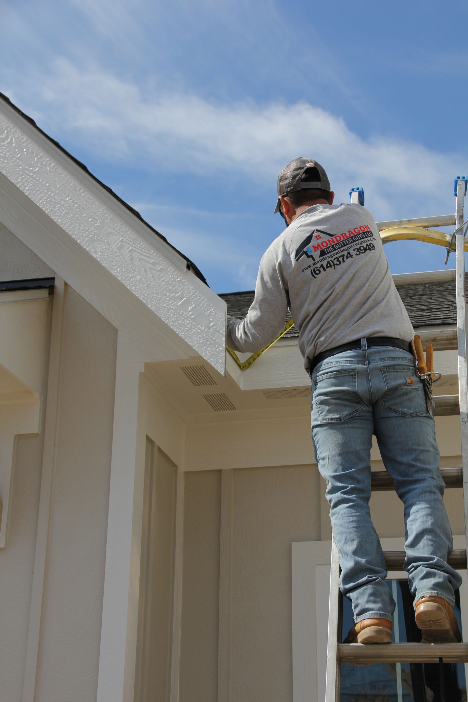 Man on ladder measuring a roof edge, blue sky, beige siding.