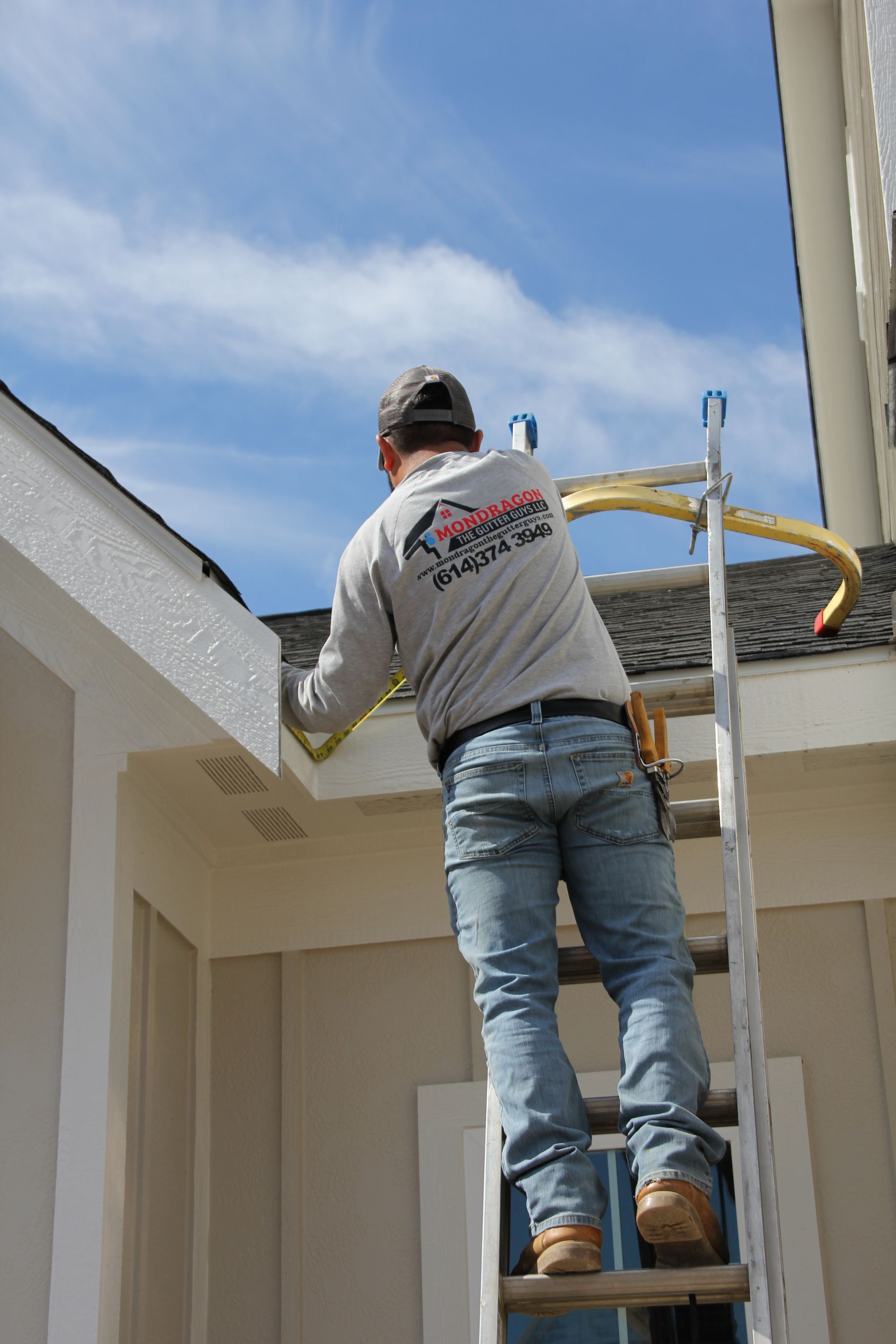 Construction worker on ladder measuring a house's gutter under a blue sky.
