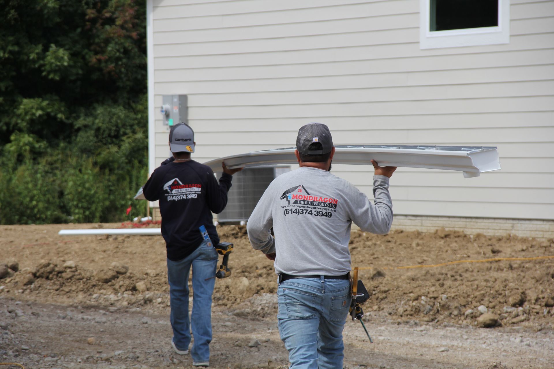 Two construction workers carrying a long metal beam near a house with tan siding.
