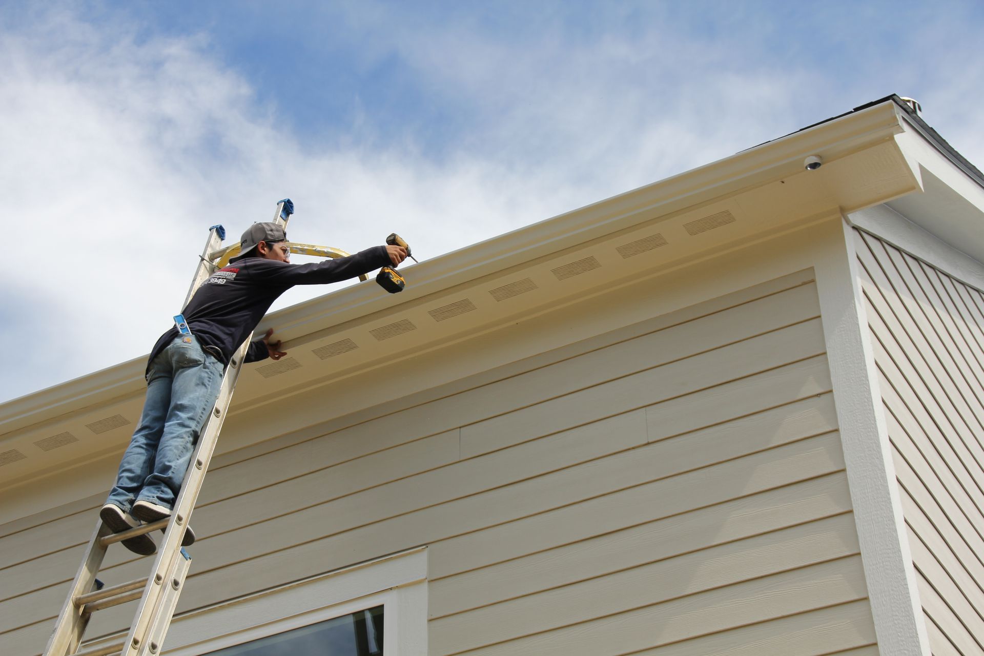 Person on a ladder drilling into the beige soffit of a light-colored house against a cloudy sky.