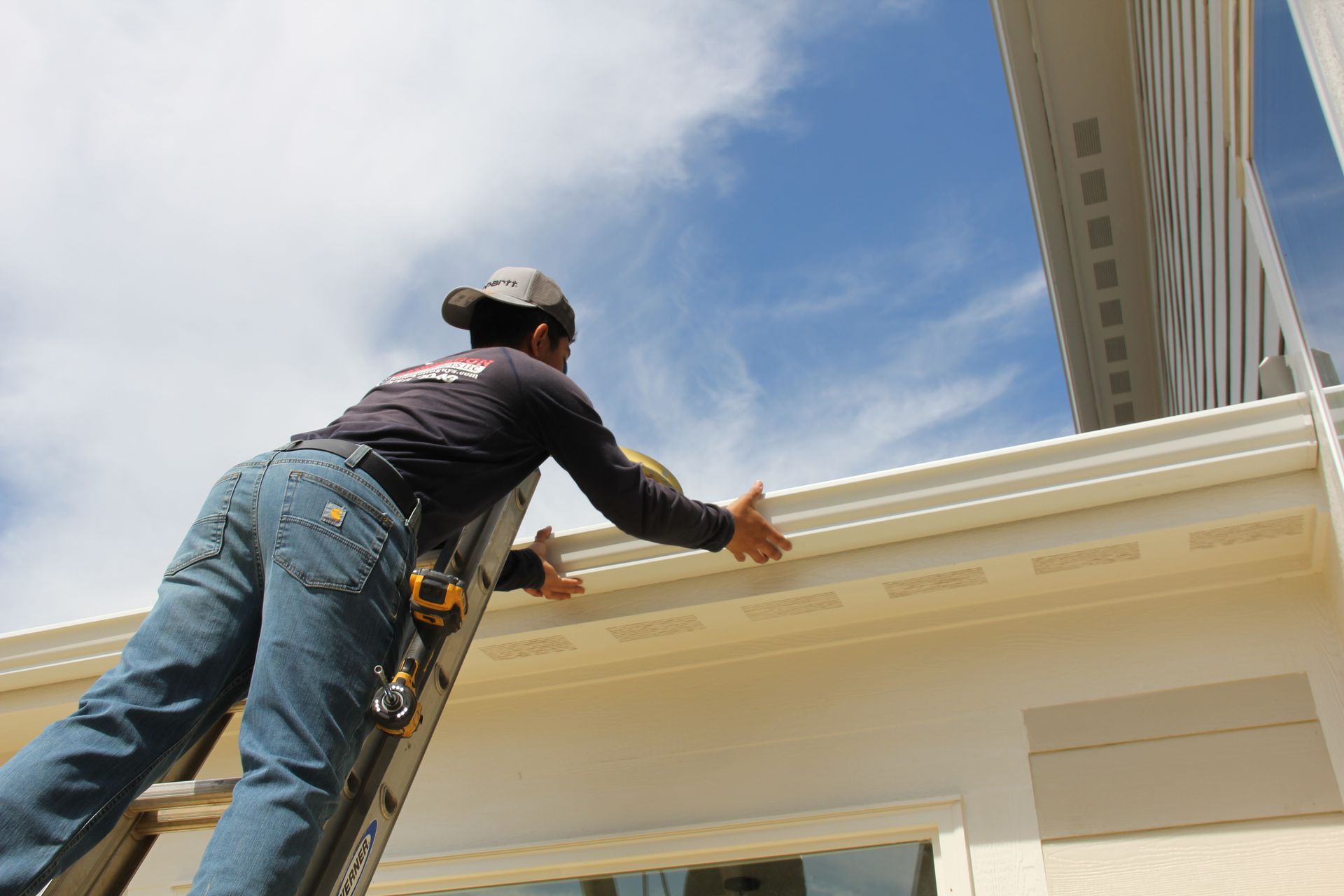 Man on ladder installing a white gutter on a house, under a blue sky.