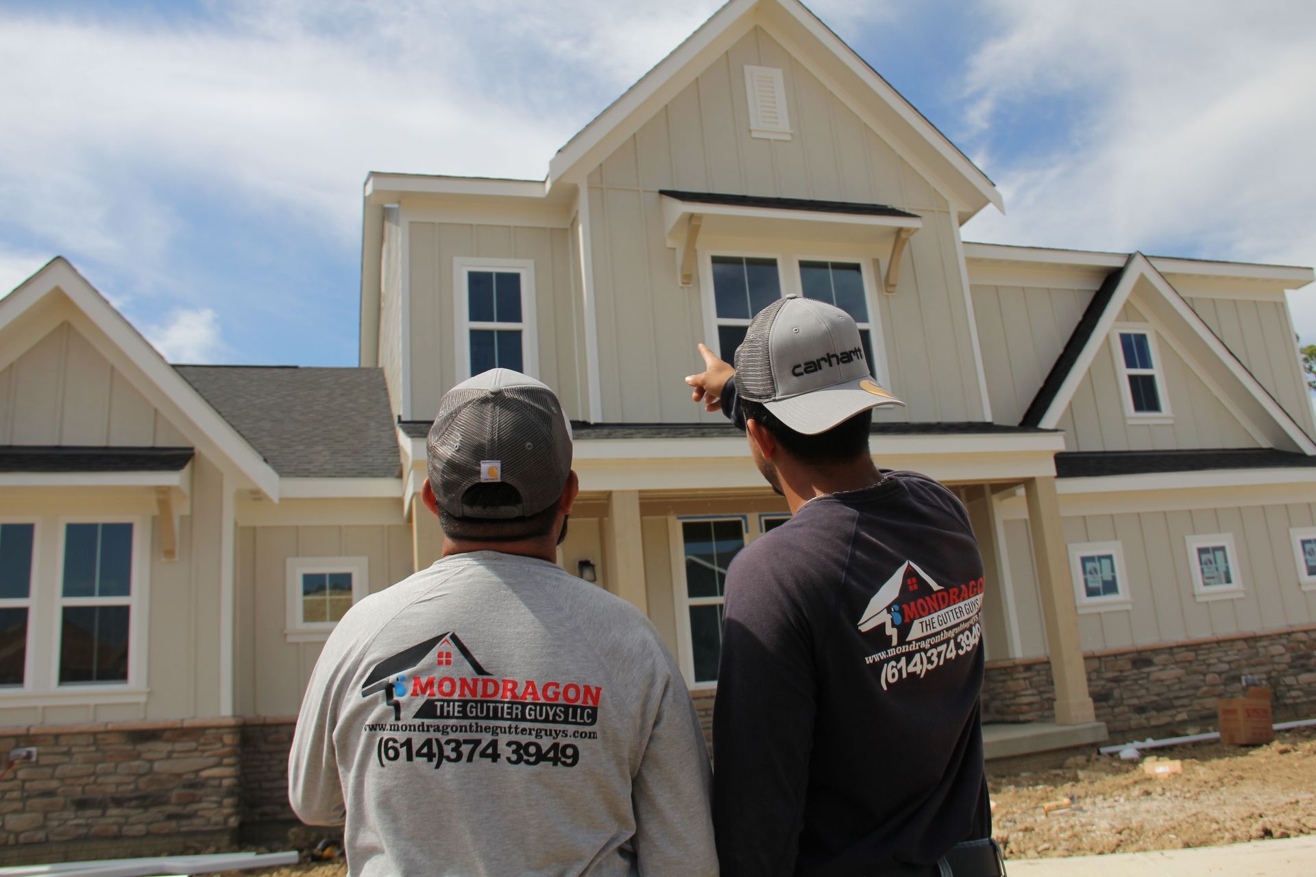 Two men in front of a light beige house; one points to the second story, discussing construction.