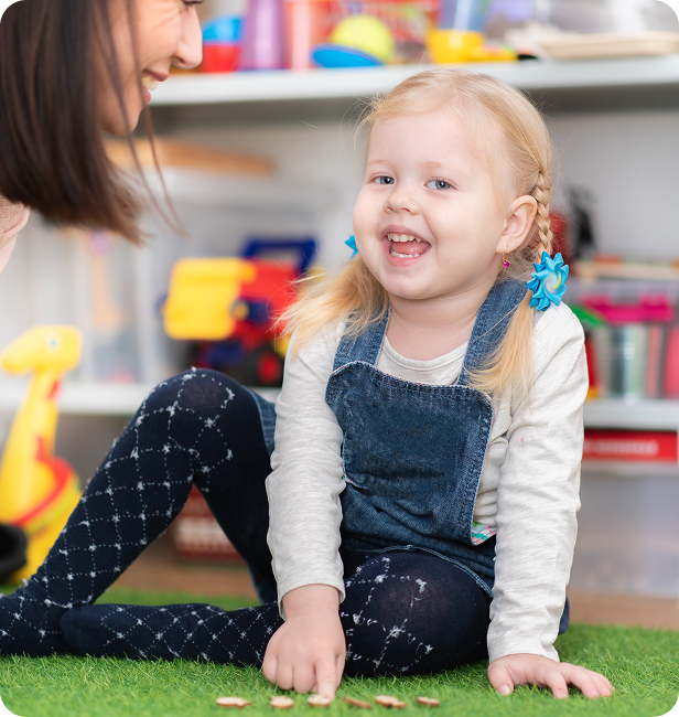 Child smiles, pointing at coins on green mat with an adult nearby.