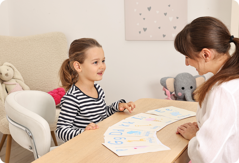 A young girl speaks to an adult, cards with numbers on the table in front of them, in a light-filled room.
