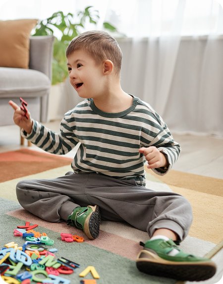 Boy with Down syndrome sitting on a rug, playing with colorful letters, smiling.