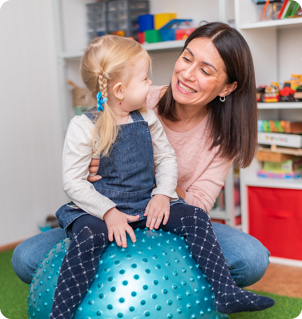 Woman and child seated together on blue exercise ball, smiling at each other.