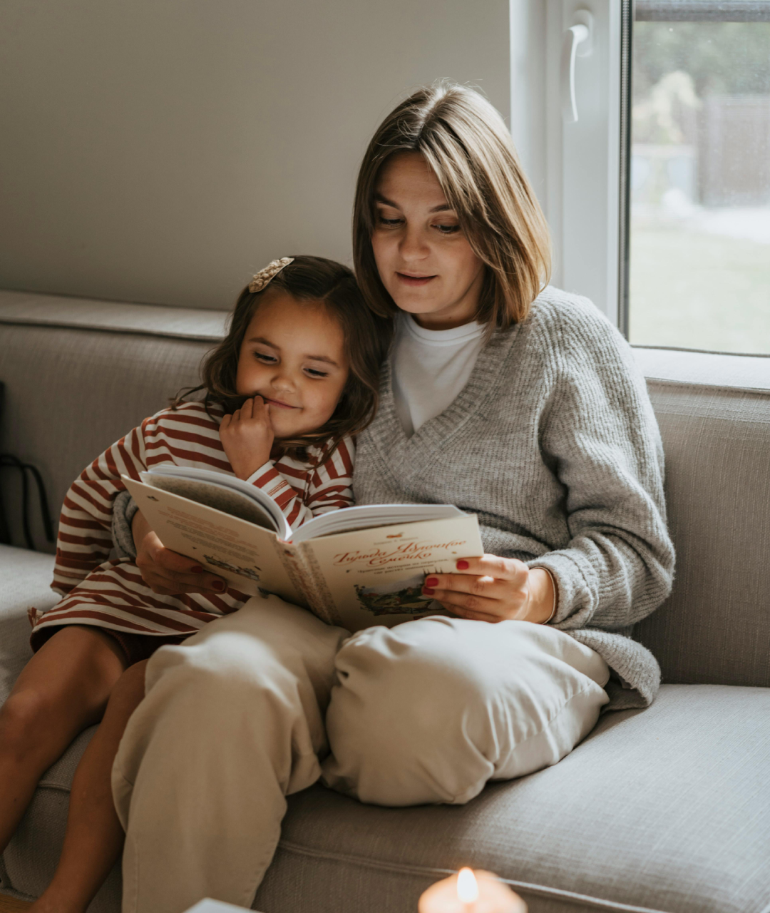 Woman reading to child on a couch, holding a book, cozy indoor setting.