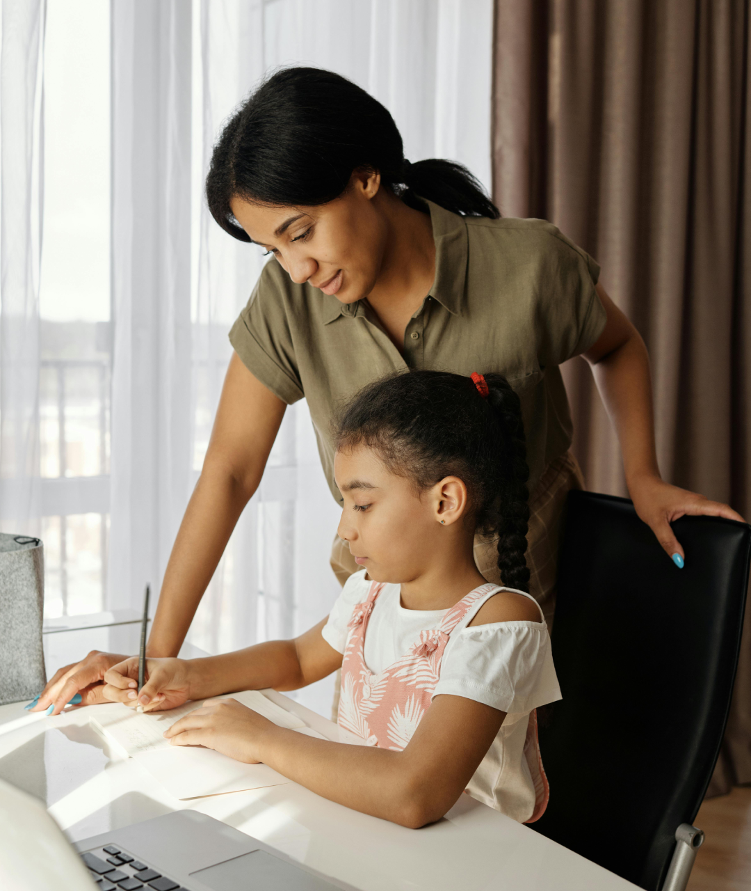 Woman helps a child with writing at a desk, a laptop visible. Window and curtains in the background.