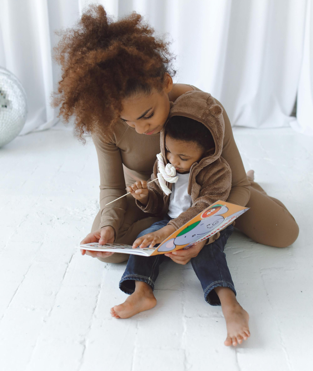 Woman reading a book with a child in a neutral-toned room. Both are looking at the book with focused attention.