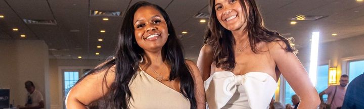Two smiling women in formal dresses pose for a photo at an indoor event with ambient lighting.