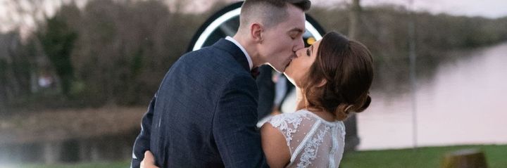A couple in wedding attire kissing in front of a ring light, set against an outdoor landscape with trees and water.