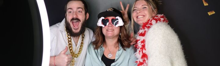 Three people posing in a photo booth, wearing fun accessories like heart-shaped glasses, a gold chain, and a red boa.
