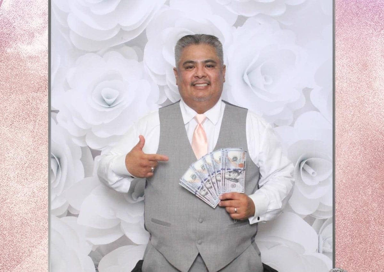 A person wearing a white shirt and gray vest smiles while holding a fan of cash in front of a white floral backdrop.