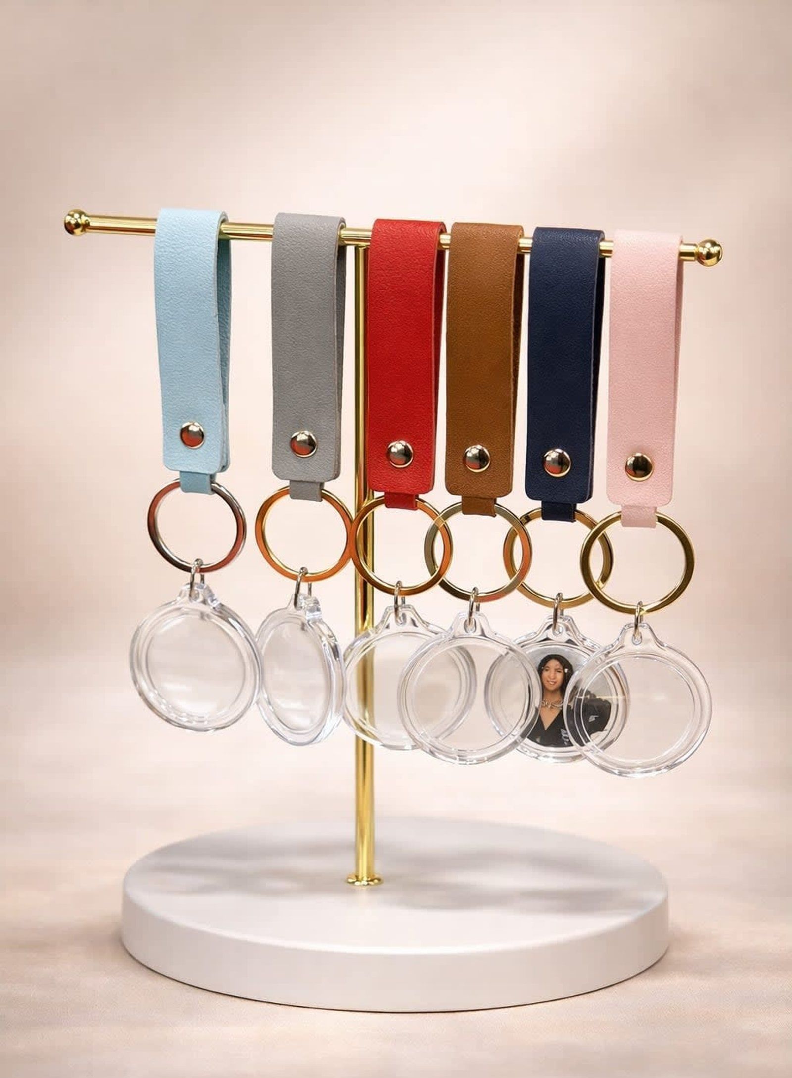 Six colorful leather strap keychains with circular clear photo frames displayed on a gold stand against a neutral background.