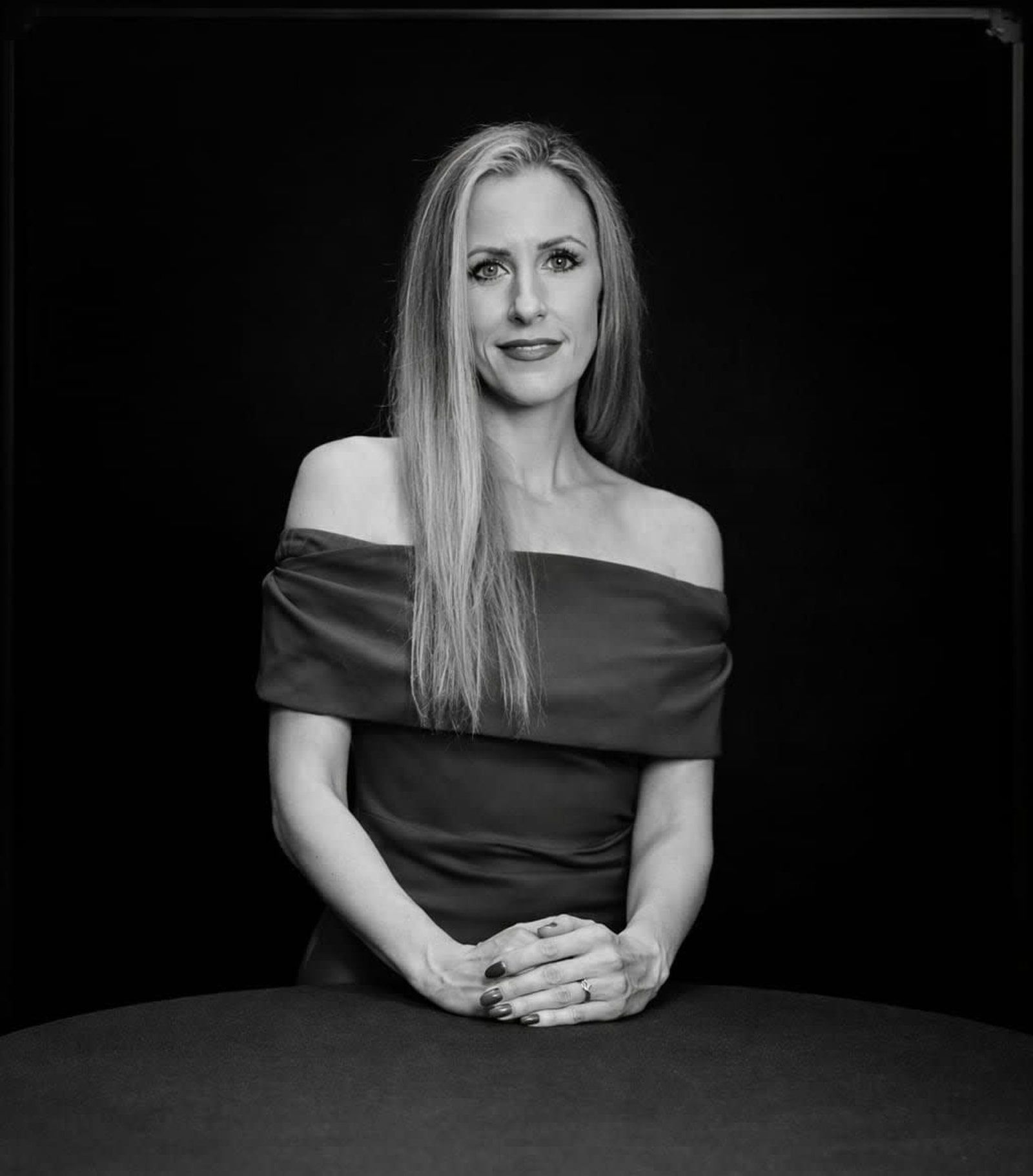 A woman with long hair wearing an off-the-shoulder top, standing with hands clasped in a studio portrait against black.