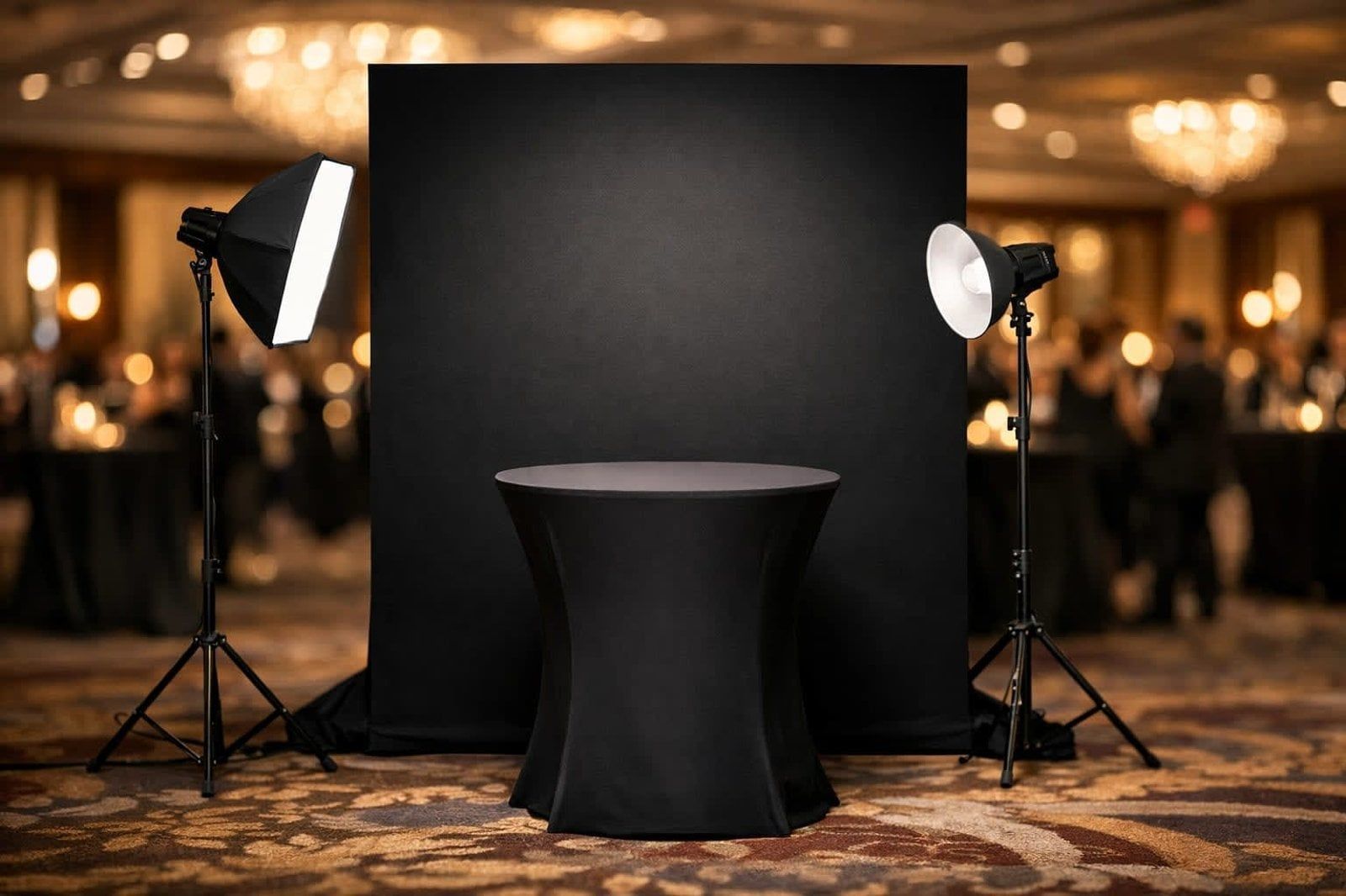 A black photo backdrop with a covered pedestal table, flanked by two studio lights in a dimly lit ballroom setting.