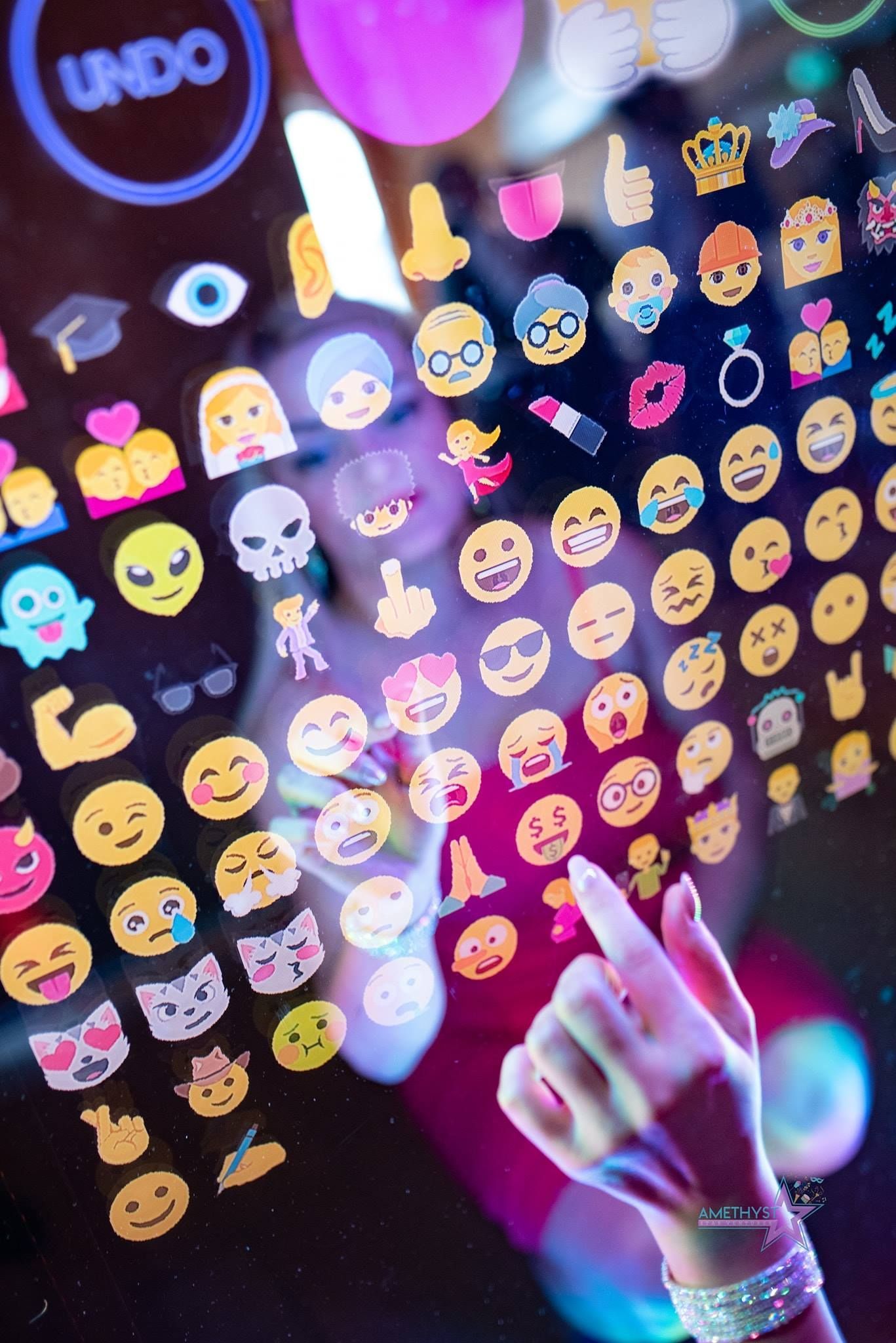 A person interacts with a touchscreen displaying a grid of various emojis, with a prominent 