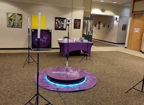 A 360-degree photo booth platform with light stands and a purple rug set up in a hallway with art on the walls.