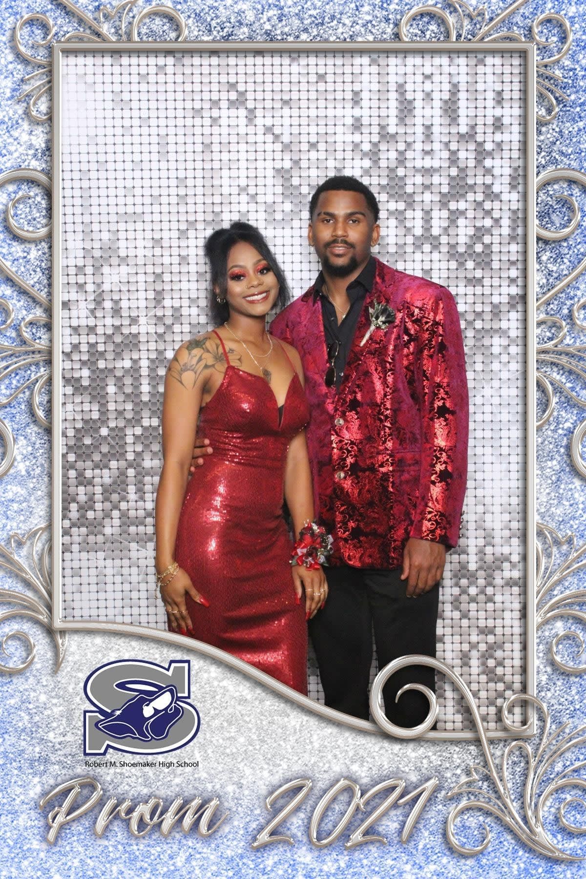 A smiling couple in red formal wear stands against a sparkly silver backdrop in a decorative Prom 2021 photo frame.