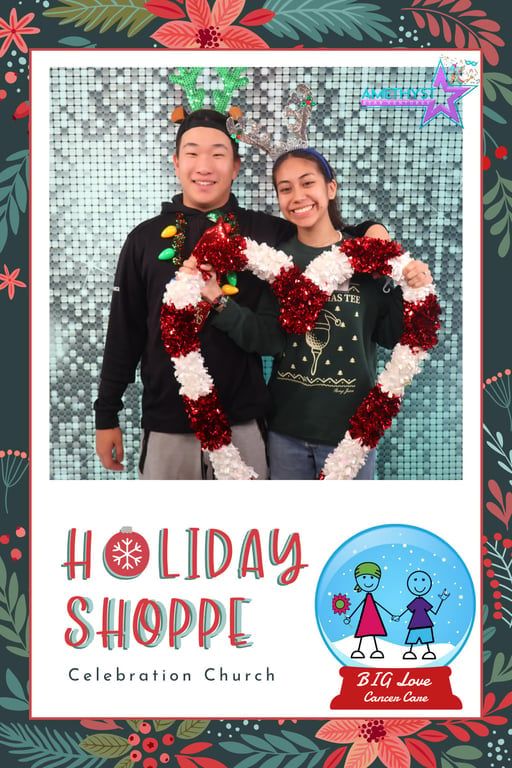 Two people pose together holding a large heart-shaped garland in front of a holiday-themed