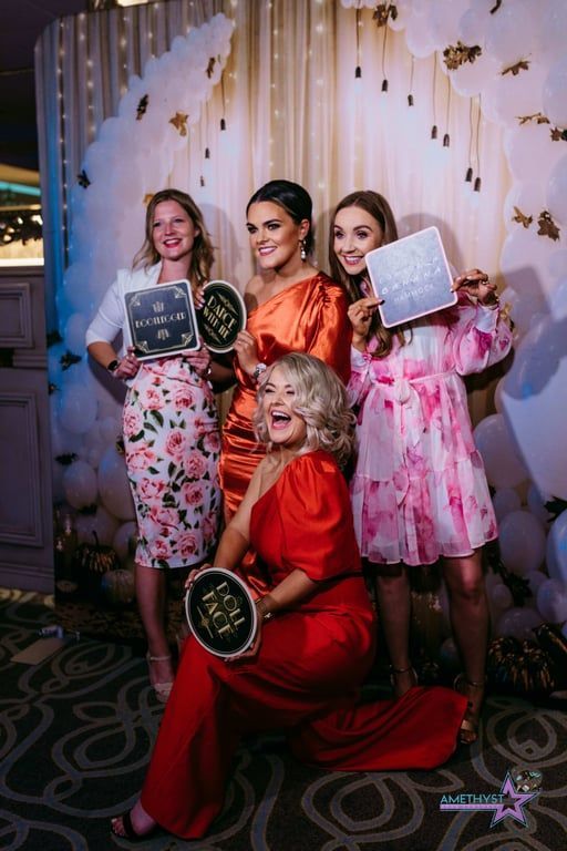 Four women pose in front of a decorative backdrop, smiling and holding event props at a celebration.