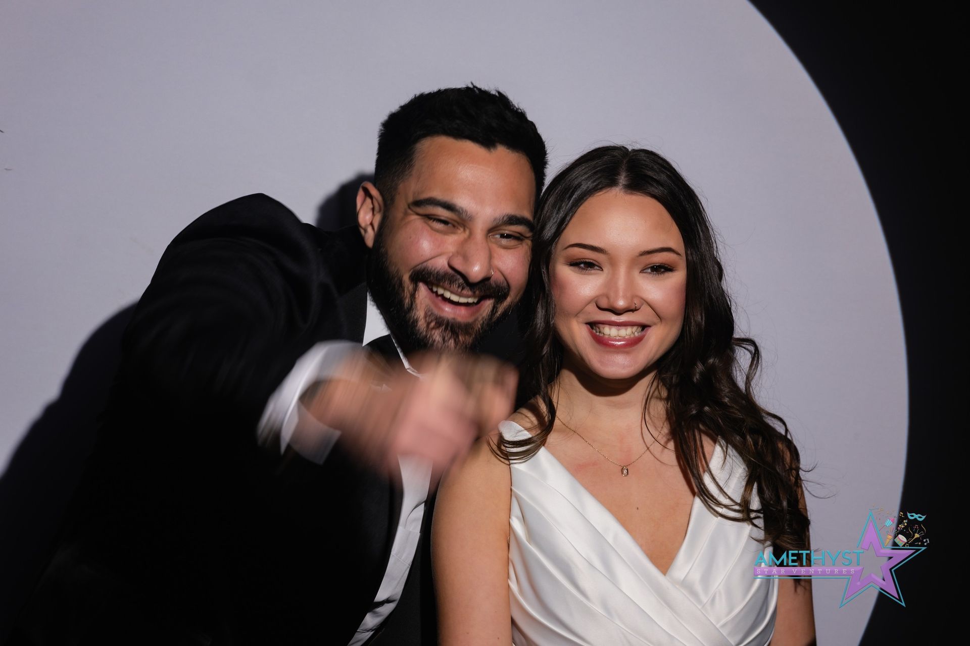 A smiling man pointing forward stands next to a woman in a white dress in front of a circular white backdrop.