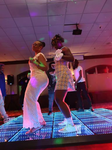 Two people dance on an illuminated LED dance floor in a dark room with purple lighting.