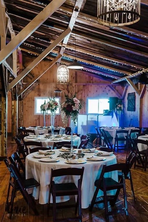 A rustic barn wedding reception space featuring round tables with white tablecloths, floral centerpieces, and dark chairs.
