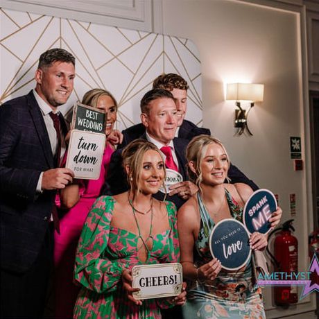 Six people smile at a photo booth, holding signs that say 