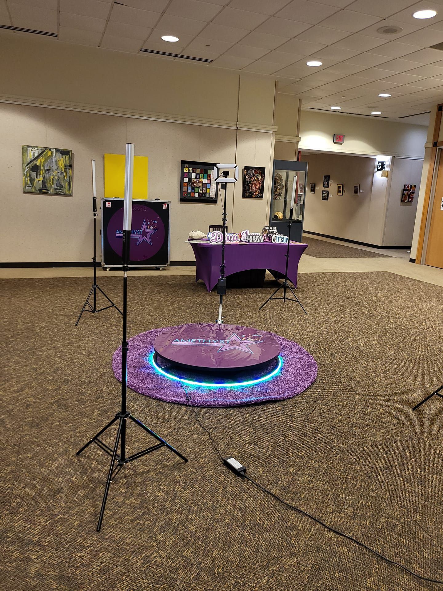A 360-degree photo booth setup on a purple rug, surrounded by lighting stands, in a carpeted indoor hallway.