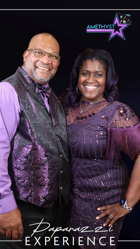 A smiling man and woman pose together wearing matching purple formal attire against a black background with event branding.