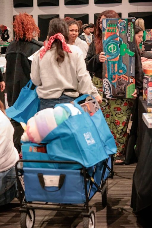A person pulls a blue wagon full of bags and a plush toy while another person holds a large Nerf toy box indoors.