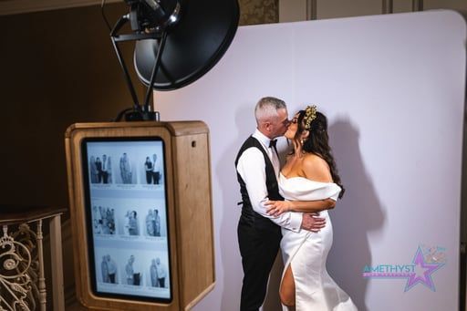 A bride and groom kissing in front of a white backdrop next to a wooden photo booth at a wedding.