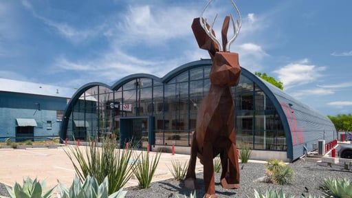 A large, metallic jackalope sculpture stands before a curved, glass-fronted building under a bright blue sky.