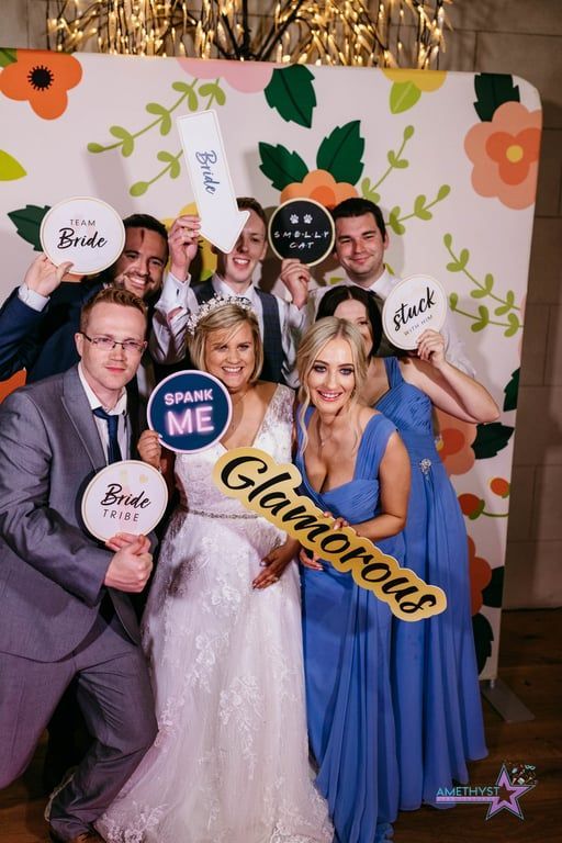 A bridal party poses with fun props in front of a floral backdrop at a wedding reception.