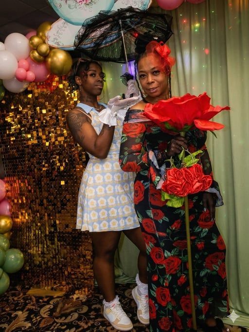 Two people in costumes, one holding a parasol and the other a large decorative rose, posing in front of a party backdrop.